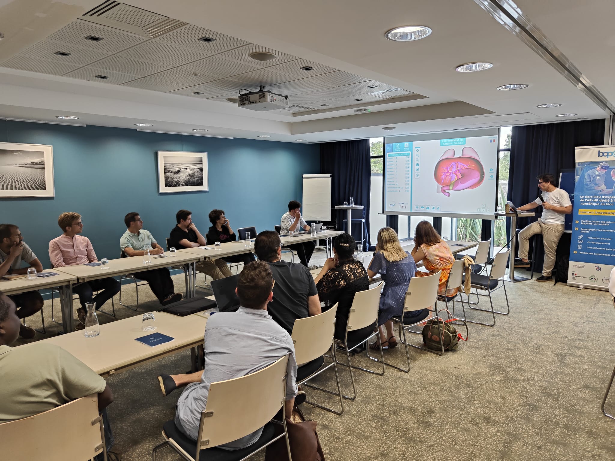 A speaker presents a 3D anatomical model during a Surgical Innovation Weekend workshop, in front of a seated audience.